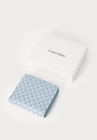 Calvin Klein lommebok i lyseblå med et mønster av mørkeblå sammenflettede former, glidelås lukking og glatt tekstur, plassert ved siden av en hvit boks.