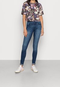 Camisa corta con estampado floral en púrpura, rosa y beige; combinada con jeans ajustados de color azul oscuro y zapatillas blancas con acentos en tonos pastel.