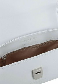 L'intérieur d'un sac à main en cuir blanc montre une poche zippée et un logo embossé "Lancaster Paris fabriqué en France". Texture lisse, forme rectangulaire.