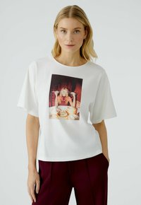 Witte katoenen t-shirt met een ronde hals en losse korte mouwen, voorzien van een kleurrijke grafische print van een vrouw met voedsel.