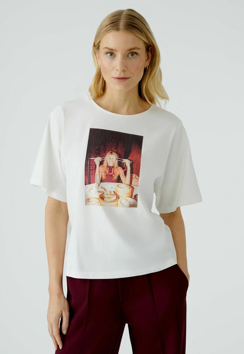 Witte katoenen t-shirt met een ronde hals en losse korte mouwen, voorzien van een kleurrijke grafische print van een vrouw met voedsel.