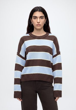 Jeune femme aux longs cheveux foncés porte un pull rayé marron et bleu clair ainsi qu'un pantalon foncé, debout devant un fond clair uni.