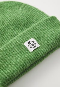 Bonnet en tricot vert avec une texture côtelée, arborant une étiquette blanche avec un logo noir circulaire. Matériau doux et design à revers.