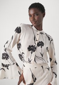 Blusa chiara in tessuto setoso, con stampa floreale nera. Maniche lunghe con polsini svasati e chiusura frontale a bottoni.