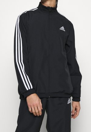 Personne portant un survêtement noir Adidas avec des bandes blanches sur les manches et les pantalons, debout devant un fond clair uni.