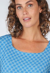 Blaue gemusterte Bluse mit rundem Ausschnitt und wellenförmigen geometrischen Designs. Der Stoff scheint leicht und mit einer glatten Textur zu sein.