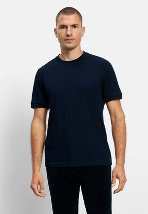 Uomo con capelli corti che indossa una maglietta blu scuro tinta unita a maniche corte e pantaloni neri, in piedi davanti a uno sfondo chiaro e uniforme.