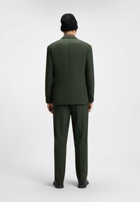 Grön kostym med skräddarsydd design, som har en enkel slits, slim fit-byxor och en svart stickad mössa. Slät textur, modern silhuett.