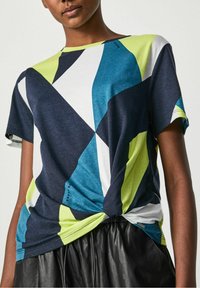 Krátký rukáv, vícebarevné tričko s geometrickým vzorem v námořnické modré, tyrkysové, bílé a limetkově zelené; uzel na lemu.