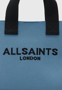 Niebieska torba tote o teksturowanej powierzchni z czarnymi nylonowymi rączkami, na której wyeksponowane jest logo "ALLSAINTS LONDON" w wyraźnych czarnych literach.