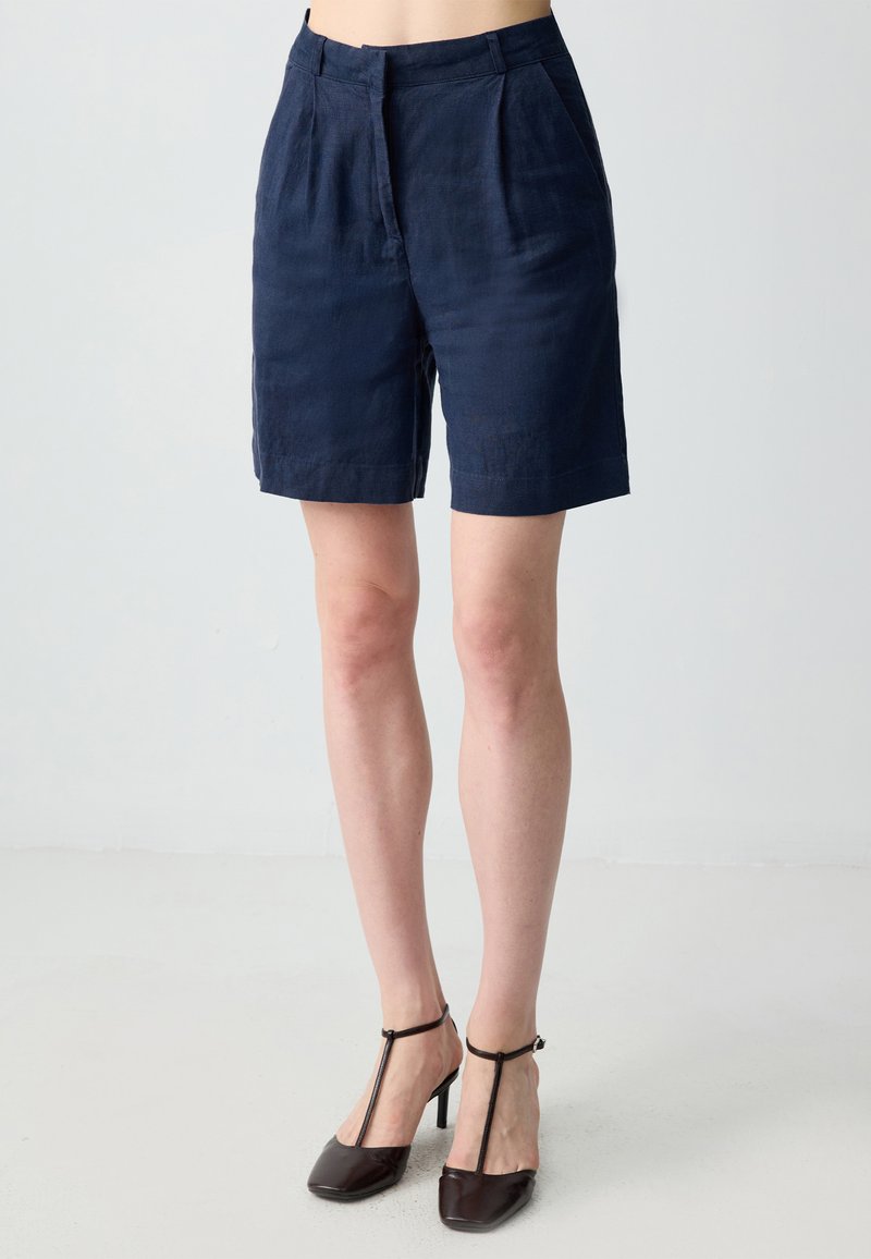 Pantaloni corti blu navy realizzati in un tessuto leggero, con pieghe frontali, una vestibilità su misura e un taglio dritto, che arriva a metà coscia.