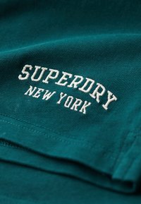 Teal Baumwoll-T-Shirt mit weißem gesticktem Text "SUPERDRY NEW YORK", mit glatter Textur und geraden Nahtdetails.
