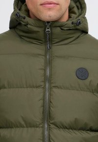 Man wearing an olive green quilted jacket with a high collar and hood, front zipper, and a small round black logo on the chest.