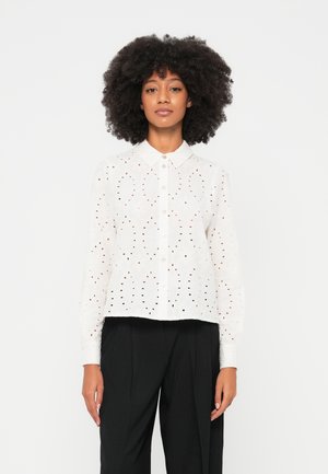 Chemise blanche à manches longues avec col, présentant des motifs d"embroidery ajourée complexes et un design court, portée sur un pantalon noir taille haute.