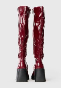 Knee-high, glossy burgundy boots with a fitted design, textured black block heels, and rear zipper closures.
