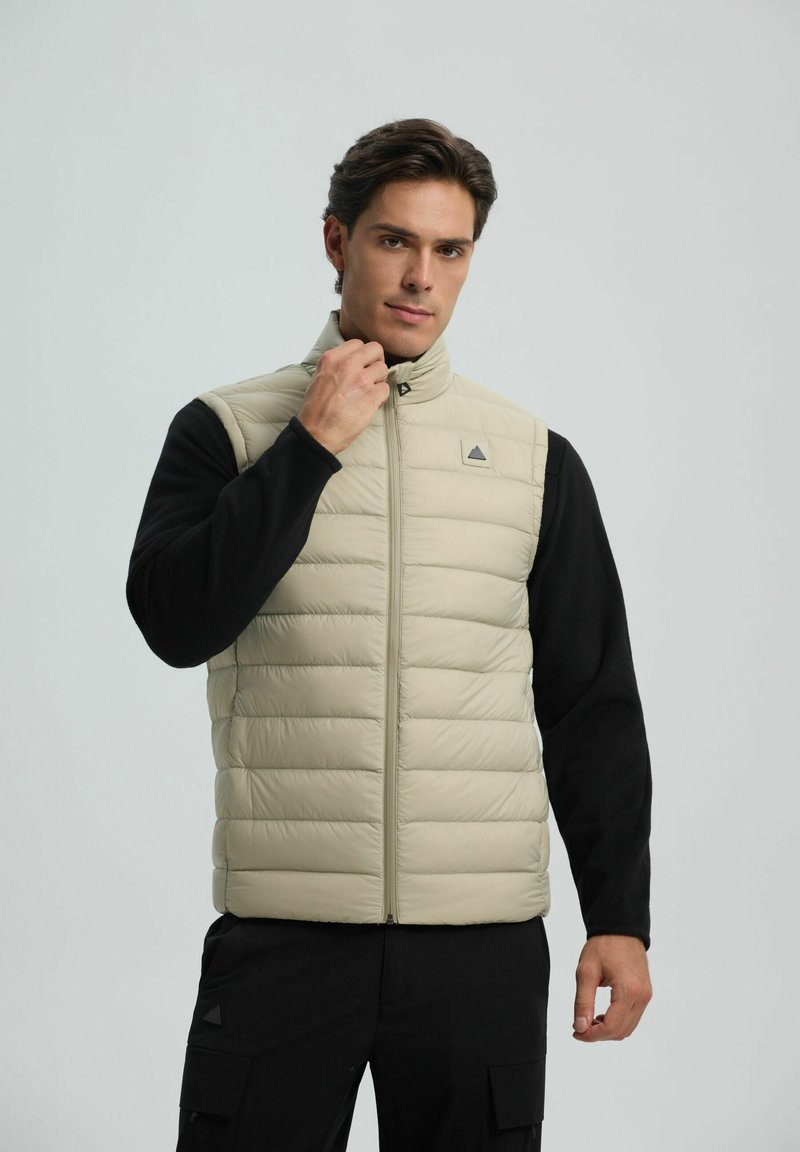 Uomo che indossa un gilet trapuntato beige sopra una maglietta a maniche lunghe nera, mentre sistema il colletto del gilet, in piedi contro uno sfondo grigio chiaro.