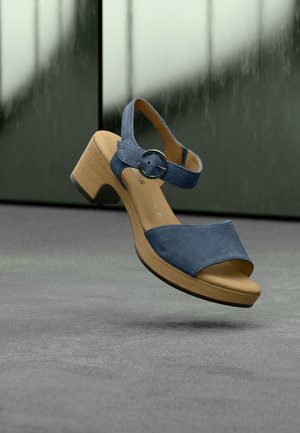 Sandale à talon en daim bleu avec bout ouvert, bride de cheville avec une boucle circulaire et texture en bois sur le talon.