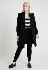 Blonda sieviete, tērpta melnā garā mētelī, melnbaltā svītrainā krekla, melnās biksēs un melnās kurpēs, stāv pretī vienkāršai gaišai fona.