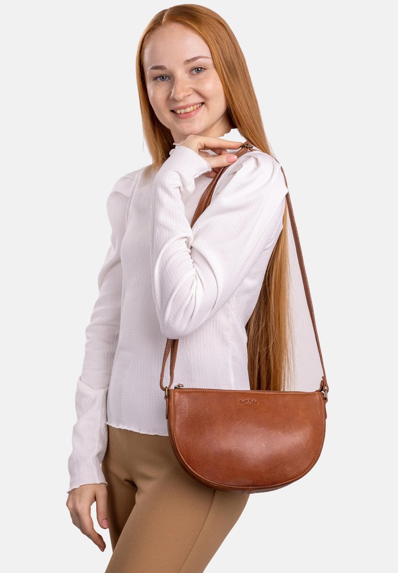 Braune Leder-Umhängetasche mit geschwungener Form, glatter Textur und verstellbarem Riemen, über der Schulter getragen zu einem lässigen Outfit.