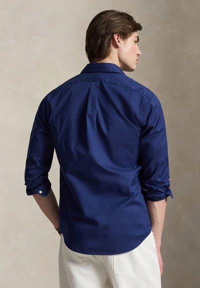 Άνδρας που κοιτάζει προς τα πίσω, φοράει μπλε ναυτικό πουκάμισο με μακριά μανίκια, τα οποία είναι σηκωμένα, και λευκό παντελόνι, σε ουδέτερο φόντο.