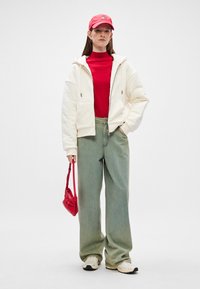 Weißer Reißverschluss-Hoodie, roter Rollkragenpullover, hellgrüne Schlagjeans, rote Handtasche und weiße Sneaker. Rote Kappe mit Logo.