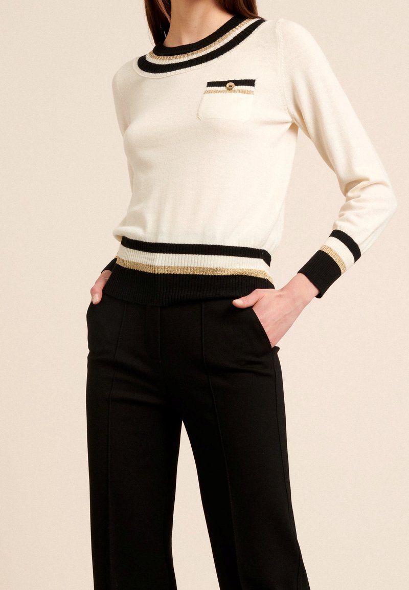 Maglione bianco con dettagli neri e dorati, colletto e polsini a coste, dotato di una piccola tasca sul petto. Abbinato a pantaloni neri tagliati.