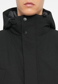 Zwarte hooded jack van een textured stof, met een hoge kraag, twee zwarte drukknoopjes en een verstelbare trekkoord. Glad binnenvoegsel.
