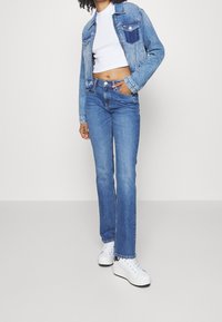 Chaqueta de mezclilla con un lavado en azul y ligero desgaste, combinada con vaqueros de talle alto en azul y zapatillas blancas, de pie sobre un fondo claro.