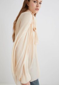 Blusa pesca pallido con maniche lunghe e fluide e un fiocco al collo. Realizzata in tessuto leggero con una texture liscia e morbida.