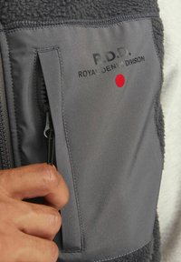 Grijze fleece vest met een gladde stofpaneel, ritszak, zwart gedrukt logo en rode stipdetail op de borst, hand dat de rits vasthoudt.