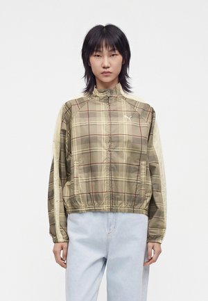 Donna con capelli lisci e scuri che indossa una giacca beige a quadri con cerniera e jeans azzurri chiari, in piedi davanti a uno sfondo bianco semplice.