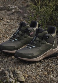 Pair of green and black Terrex hiking shoes with Climawarm technology placed on rocky ground with sparse vegetation.
