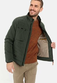 Quiltet grøn jakke med en frontlynlås, brystlomme og tan-foring. Båret over en orange struktureret sweater og khaki bukser.