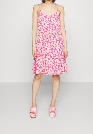 Nainen pukeutuneena vaaleanpunaiseen hihattomaan mekkoon, jossa on valkoisia päivänkakkarakuvioita, ja hänellä on jalassa väriltään vastaavat korokepohjaiset sandaalit. Hän seisoo yksivärisen taustan edessä.