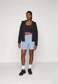 Sieviete stāv, ģērbusies melnā īsā topā, melnā kapucē, gaiši zilās velosipēdu šortos, baltās zeķēs un melnās kedās uz vienkrāsaina fona.