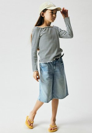 Ragazza che si sistema un cappellino beige, indossando una maglietta a maniche lunghe a righe bianco e nero, pantaloncini di jeans fino al ginocchio e infradito gialle su sfondo bianco.