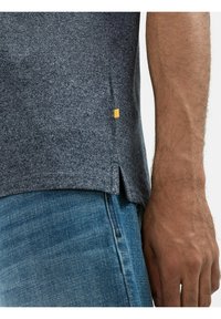 Szara koszulka z krótkim rękawem o teksturowanej tkaninie, z bocznymi rozcięciami i żółtym akcentem na brzegu, w połączeniu z niebieskimi dżinsami.