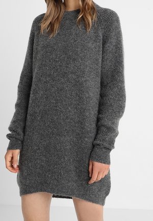 Woman wearing a long-sleeve, dark gray, fuzzy knit sweater dress standing against a plain light background.