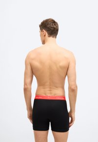 Άνδρας που στέκεται γυρισμένος πλάτη, ντυμένος με μαύρα μποξεράκια με κόκκινη ζώνη Calvin Klein, μπροστά σε απλό φωτεινό φόντο.