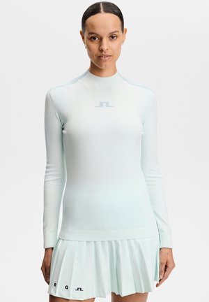 Vrouw draagt lichtblauw nauwsluitend shirt met lange mouwen en geplooide rok met subtiele logo's, staand tegen een effen witte achtergrond.