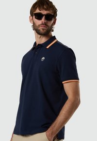 Hombre con gafas de sol oscuras y una camiseta polo azul marino con cuello y bordes de las mangas a rayas naranjas, posando frente a un fondo liso.