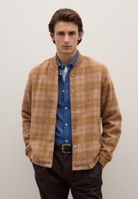 Giacca a quadri marrone in materiale morbido, con colletto a coste, bottoni a pressione e indossata sopra una camicia di denim.