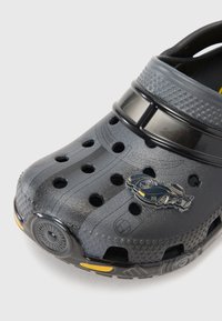 Black rubber clog with multiple ventilation holes, textured surface, and a decorative car logo, featuring a yellow accent on the sole.