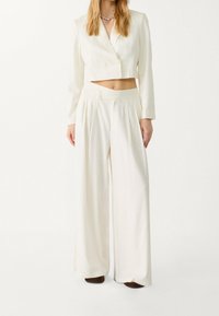 Blazer blanco corto y pantalones de pierna ancha, ambos con una textura suave y detalle de botones dobles, combinados con zapatos marrones.