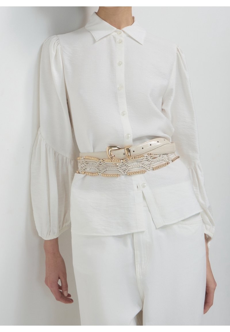 Personne portant un chemisier blanc boutonné avec des manches bouffantes et un pantalon blanc, accessoirisé d’une large ceinture beige avec des détails de chaîne dorée.