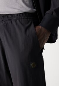 Black athletic pants with an elastic waistband, side pockets, and a textured circular logo on the left thigh. Features smooth fabric and a relaxed fit.