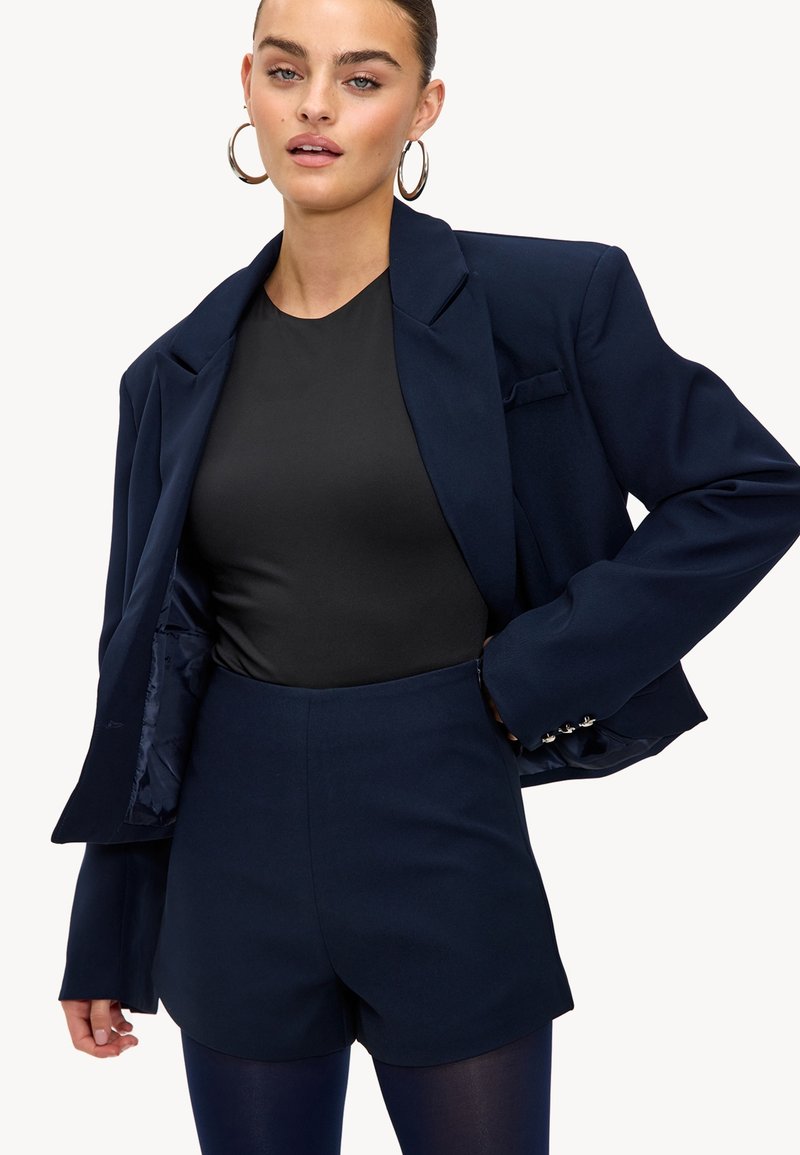 Jeune femme portant un blazer navy et un short taille haute assorti sur un haut noir, posant avec une main sur la hanche et de grandes boucles d'oreilles.