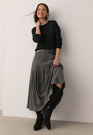Femme souriante, portant un pull noir, une jupe midi à carreaux noir et blanc, et des bottes noires hautes jusqu'aux genoux, posant contre un mur blanc.