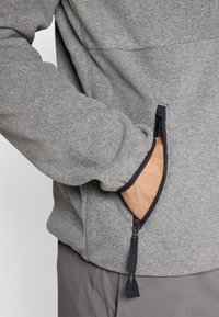 Grå fleecejakke med en svart glidelåslomme. Har en myk tekstur og avrundet design, med et nærsyn av en hånd som strekker seg inn i lommen.