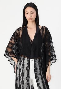 Svart spetskimono med öppen front, med geometriskt broderade mönster och vågiga kanter, lagd över en svart topp. Vita byxor synliga.